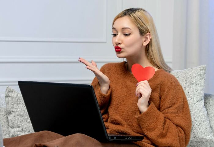 Young woman in brown sweater holding a paper heart, blowing a kiss toward laptop, exploring why guys slept with married woman online.