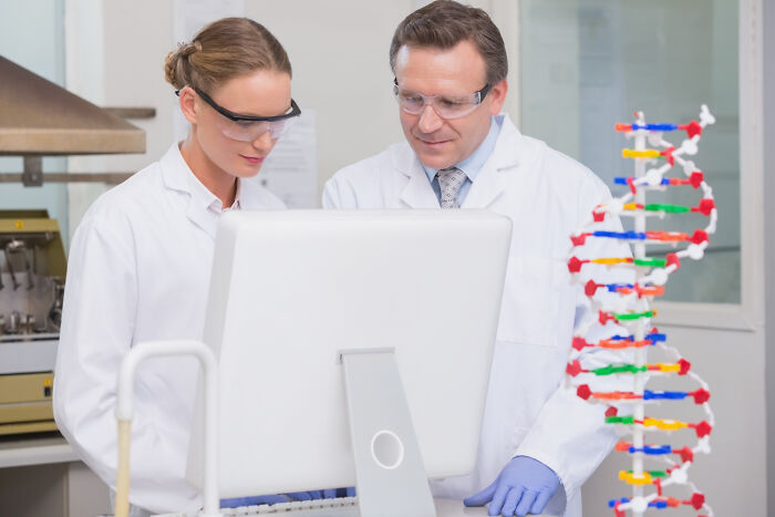 Scientists in lab coats and goggles reviewing data on a computer, with a DNA model in the foreground.