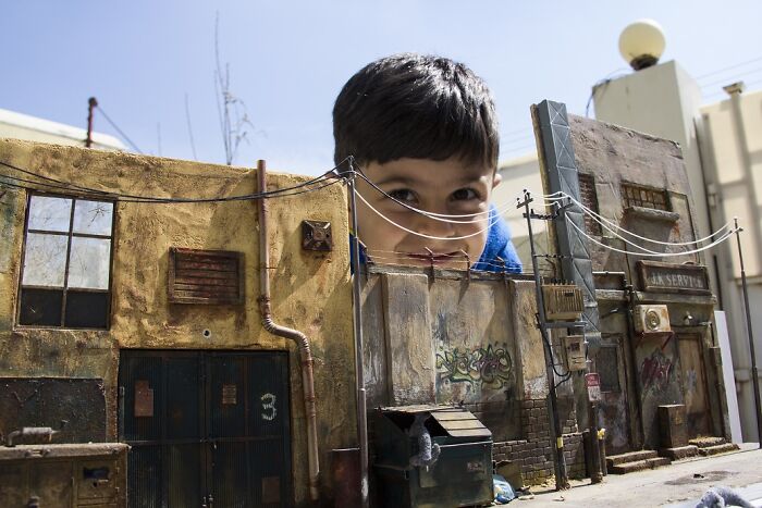 Child peeking over urban miniatures, inviting you into secret worlds.