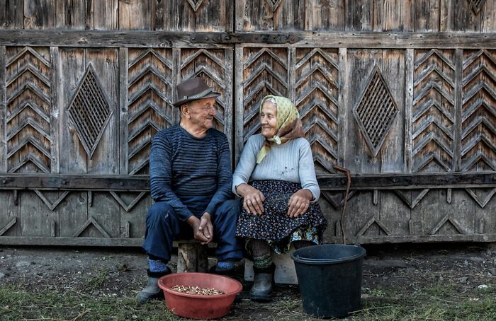 Elderly couple sitting together, smiling, in front of a rustic wooden background, highlighting the vulnerability of aging women.