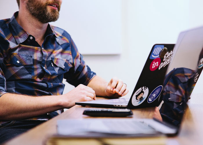 Man working on laptop covered with stickers, focused on submitting SEO deliverables in a creative office setting.