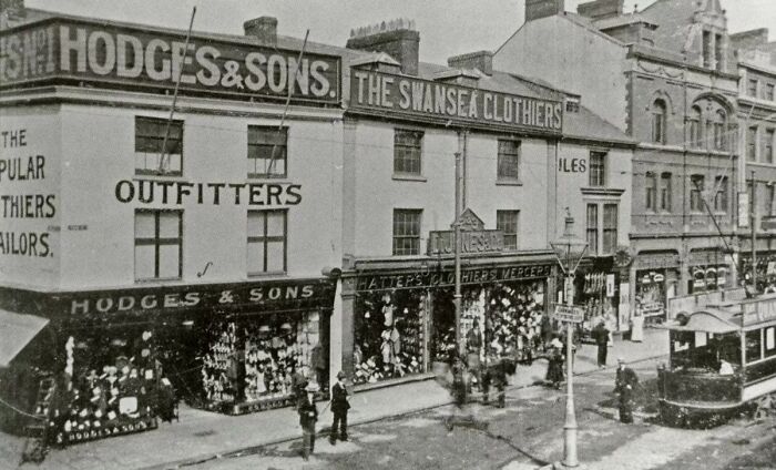 Black and white photo showing European street life 100 years ago with old shops, pedestrians, and a tram on the road.