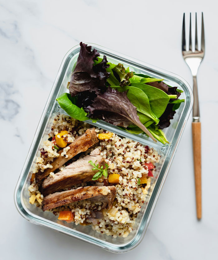 Glass meal prep container with quinoa, roasted vegetables, and salad greens on a marble surface with a fork beside it.
