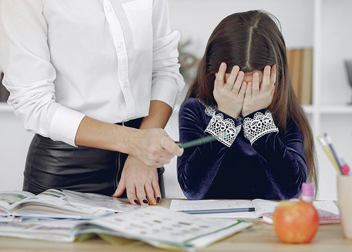 Teacher pointing at a workbook while a student hides her face in frustration during a lesson.