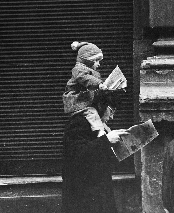 Black and white street photo of a child sitting on an adult’s shoulders, both reading newspapers outdoors.