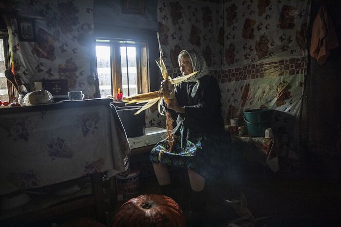 Aging woman peeling corn in a dimly lit room, conveying vulnerability and resilience.