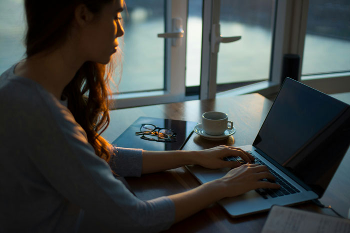 Person job hunting on laptop with coffee and glasses on wooden table by a window.
