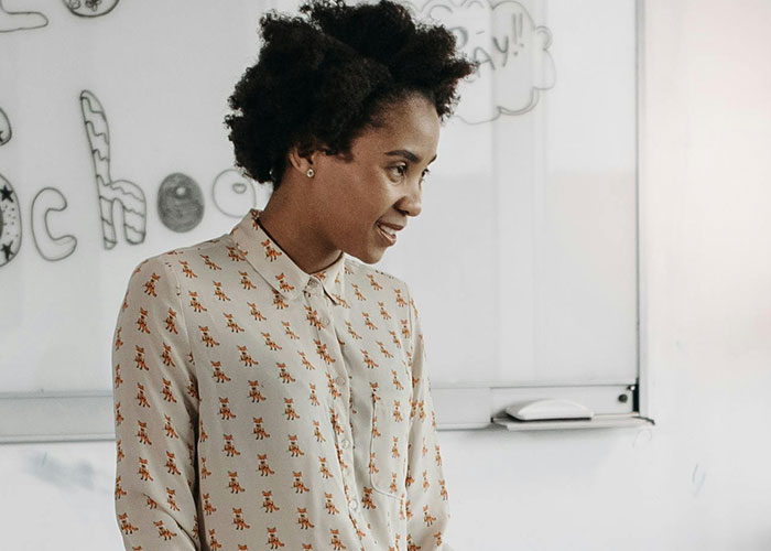 Teacher standing in front of a whiteboard with drawings, wearing a patterned shirt, engaged in teaching.
