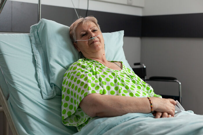 Elderly woman in a hospital bed, illustrating realities of aging, wearing a green polka dot gown with nasal cannula.