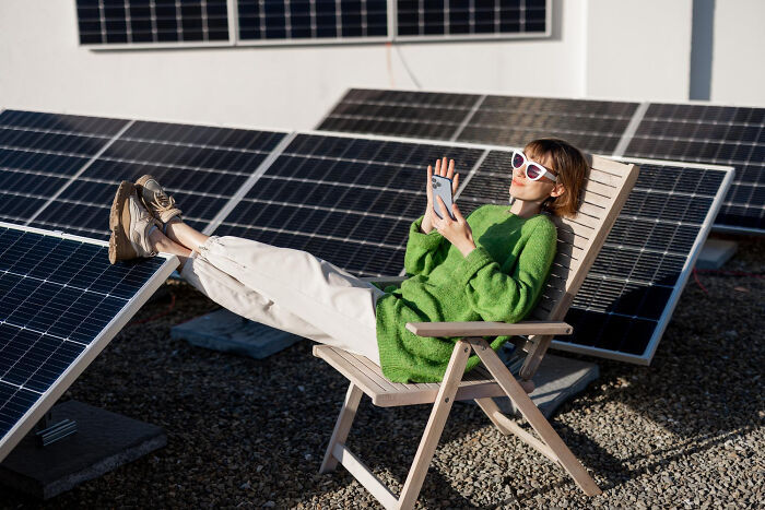 Person in green sweater confidently lounging on a chair, using a phone near solar panels.