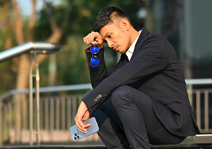 Man in suit sitting outdoors, looking contemplative, holding a phone and sunglasses, related to wedding and plus one issue.