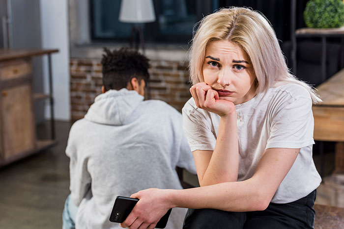 Woman looking upset with her partner turned away, holding a phone; relationship tension visible.
