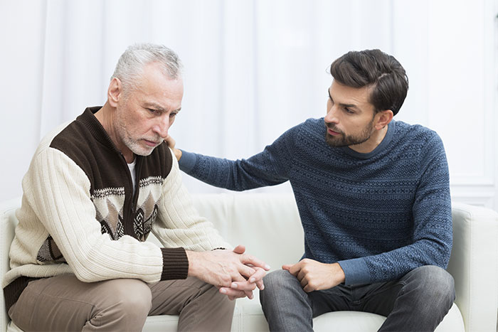 Two men sitting on a couch in conversation, one comforting the other, discussing a banned babysitting incident.
