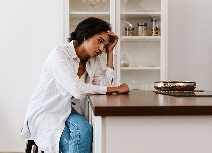 Woman sitting at kitchen counter, appearing thoughtful, wearing a white shirt and jeans, amid grocery-related dispute.