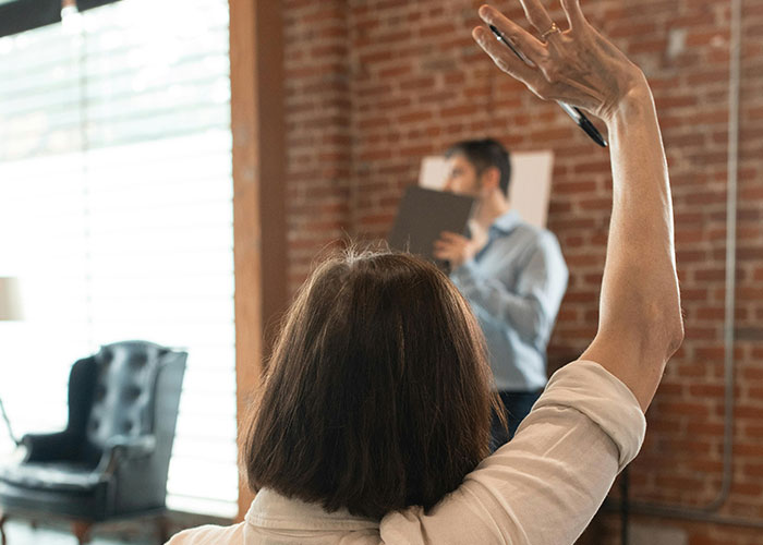 A woman raising her hand in a classroom setting with a speaker in the background, brick wall visible.
