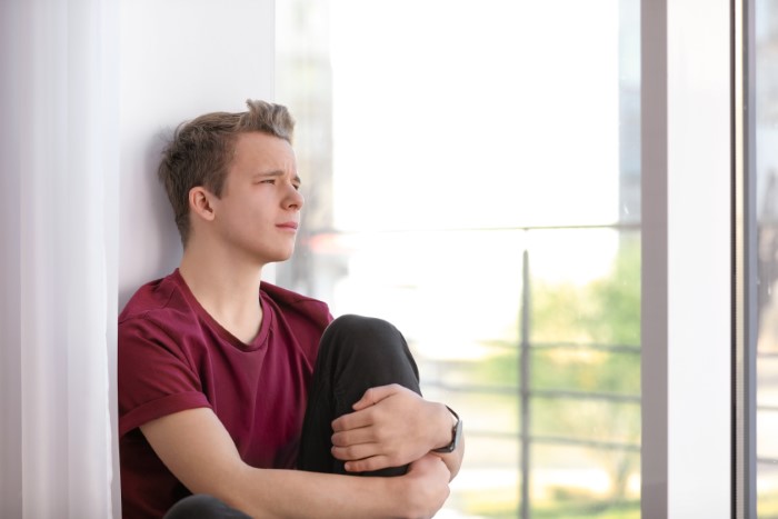 Teen sitting by a window, looking distressed, wearing a maroon shirt, contemplating harassment issues.