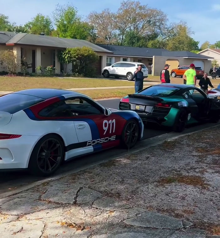 Sports cars line the street for a birthday surprise, including a Porsche 911 and a green vehicle, with people gathered nearby.