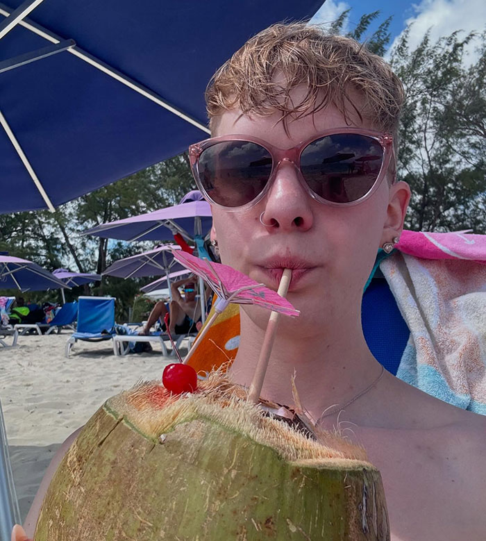 A person on the beach sipping a drink from a coconut under an umbrella, wearing pink sunglasses.