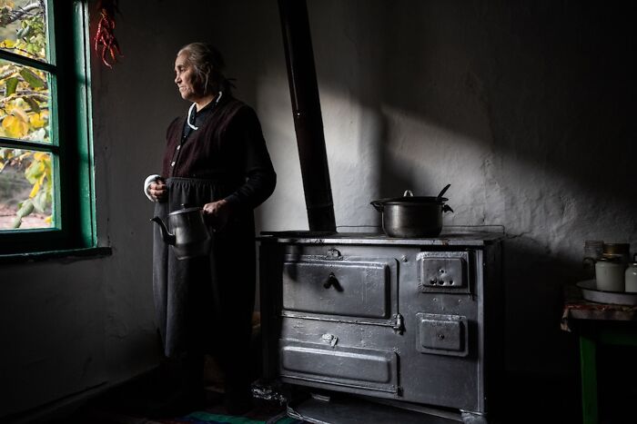 An elderly woman stands by a stove, holding a kettle, illustrating the vulnerability of aging women.
