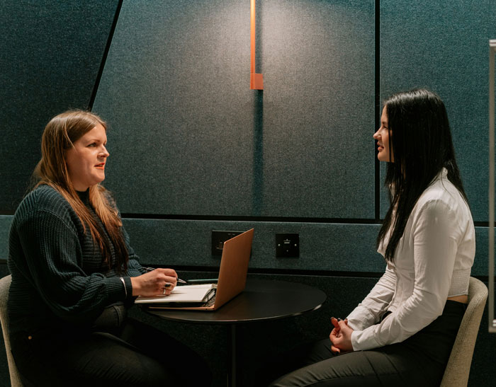 Two women in a job interview setting, one taking notes with a laptop on a small round table.