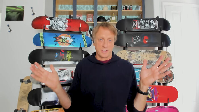 Celebrity in casual wear, gesturing in front of a wall-mounted skateboard collection, discussing experiences.