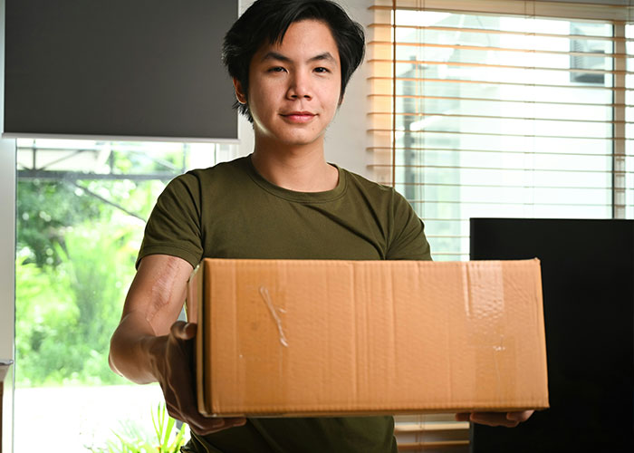 Young man in a green shirt, holding a cardboard box in a sunlit room, conveying a friendly expression.