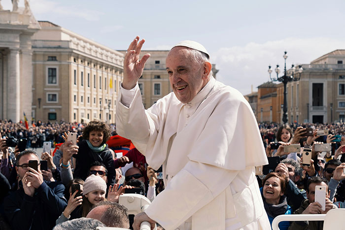 I don't know who this is, but the alt text could be: "Religious leader waving to a cheering crowd outdoors with historic buildings in the background.