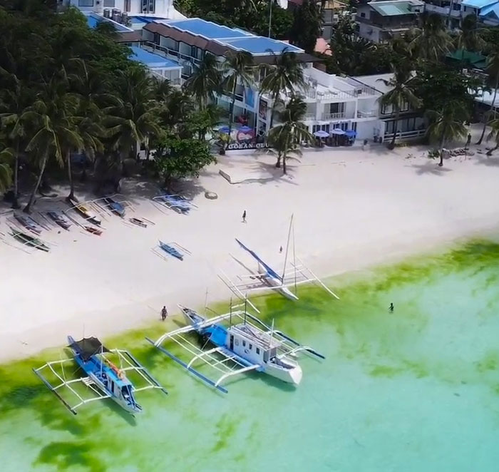 Aerial view of a beach with anchored boats and nearby buildings, highlighting a serene travel location.