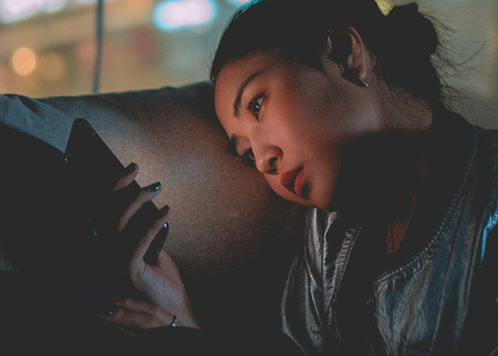 Young woman looking heartbroken while staring at her phone, reflecting on betrayal and cheating in a dimly lit room.