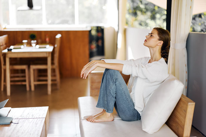 Woman sitting thoughtfully on a couch, wearing casual clothes in a bright living room.
