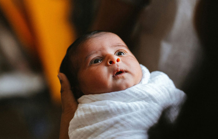 Baby wrapped in a white blanket, looking up with wide eyes, being held gently in someone's arms.