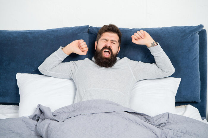 Bearded man stretching in bed, looking tired after a long night, relatable to relationship arguments and sitcom moments.