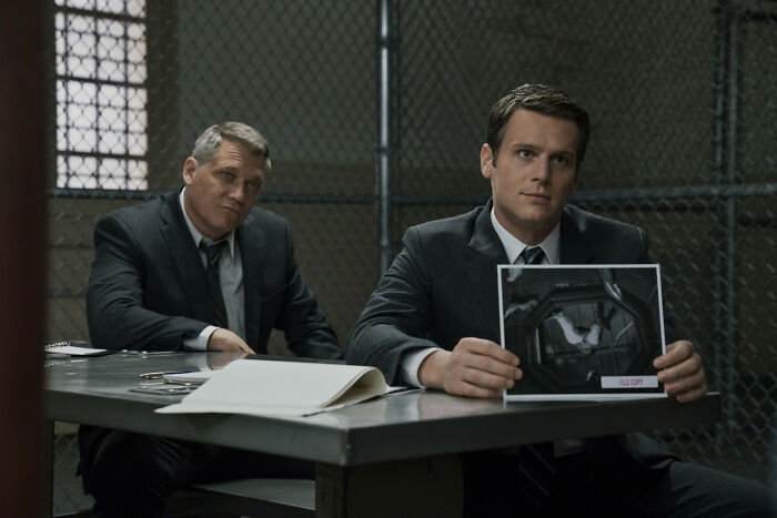 Two men in suits sit at a table in a dimly lit room, one holding a photo. Scene from a top-rated TV show.