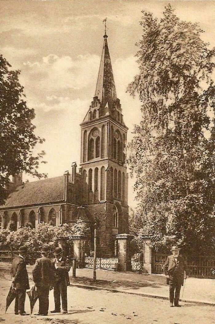Men standing near a historic church building with tall steeple in a European street scene from 100 years ago.
