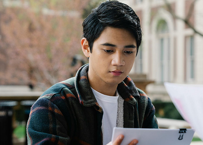 Young man in a plaid jacket reading papers outdoors, contemplating teachers and educational challenges.
