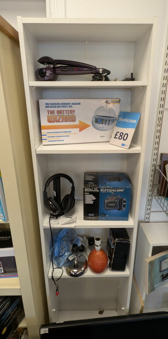 Secondhand shop shelf displaying curiosities like a Battery Wizard charger, headphones, and various gadgets.