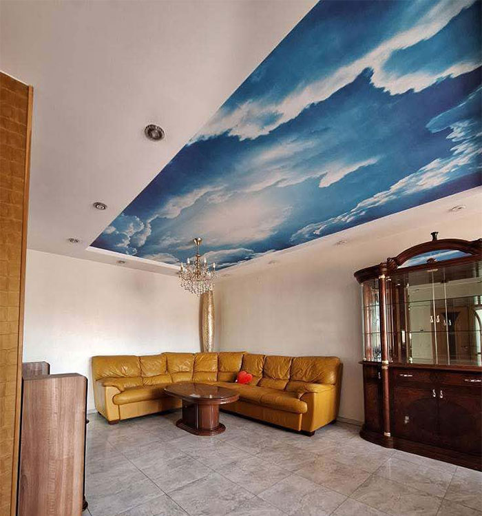 Living room with unique blue sky ceiling mural, mustard leather sectional sofa, wooden coffee table, and vintage display cabinet.