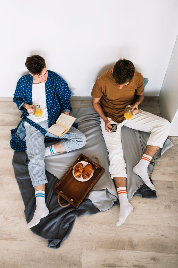 Two men sitting on the floor, enjoying croissants and orange juice, surrounded by cozy blankets.