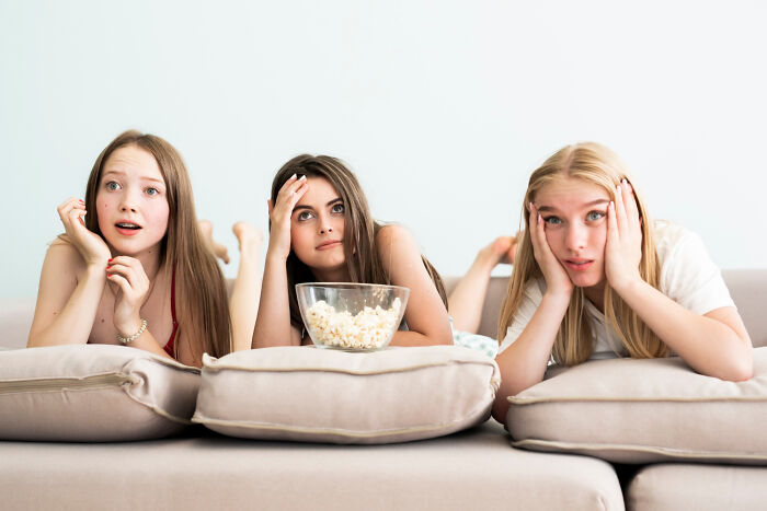 Three teens on a couch looking surprised and watching TV, with a bowl of popcorn, capturing high school incidents.