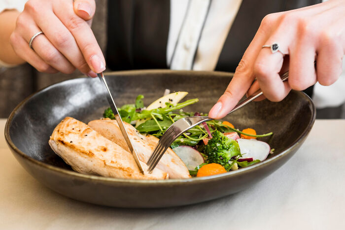 Hands cutting affordable treat of grilled chicken and fresh salad in a black bowl, highlighting fancy presentation.