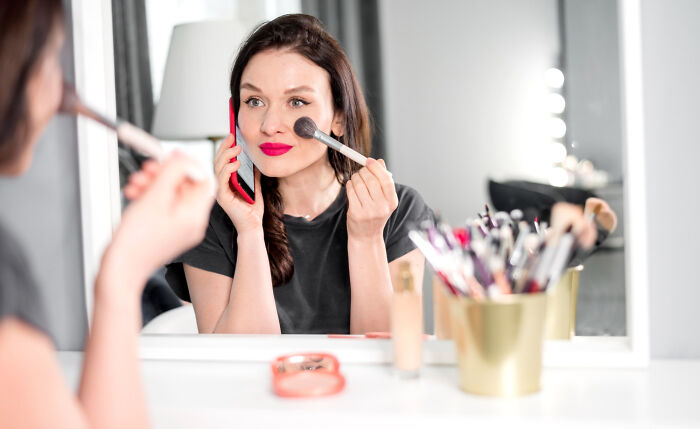 Woman applying makeup in front of a mirror, surrounded by cosmetic tools.