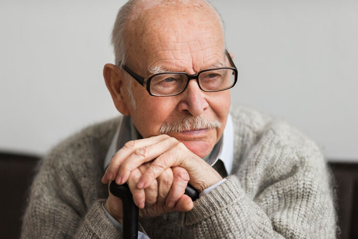 Elderly man wearing glasses, reflecting on the realities of aging with hands resting on a cane.