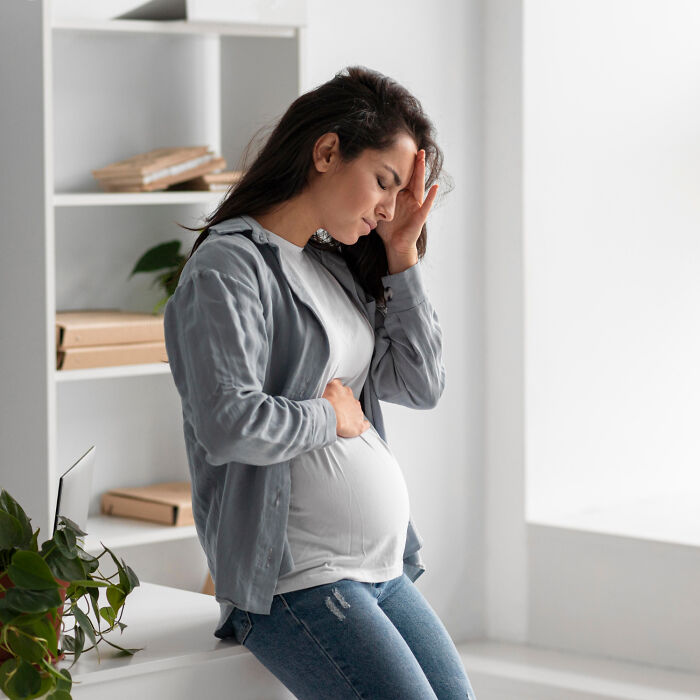 Pregnant woman in casual attire holding head and stomach, appearing thoughtful, possibly trusting her gut instinct.