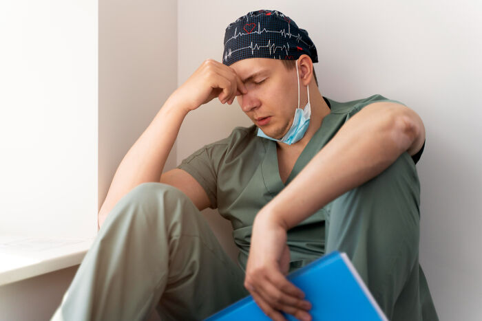 Doctor in scrubs sitting with a tired expression, holding a blue folder, illustrating stressful questions from friends.