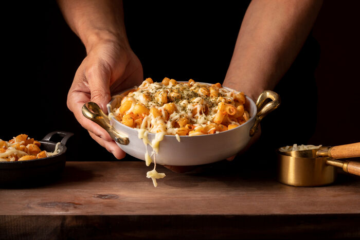 Hands serving cheesy pasta in a bowl, with golden handles, on a wooden table.