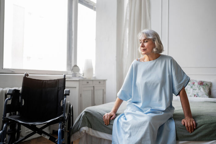 Elderly woman in blue dress sitting on bed, looking at empty wheelchair, reflecting on regretted picking favorites.