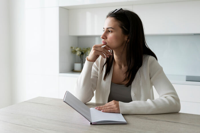 A woman in a kitchen thinking deeply, holding a notebook, reflecting on parenting and picking favorites.