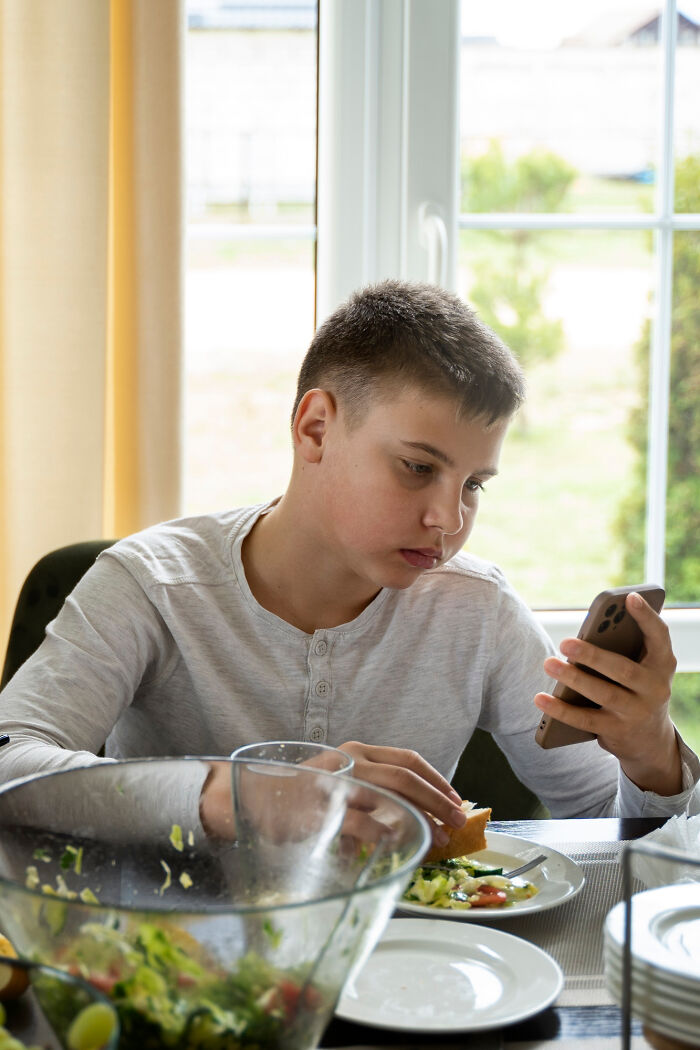 Teen boy at dining table engrossed in phone, surrounded by salad, embodying bizarre house rule vibes.