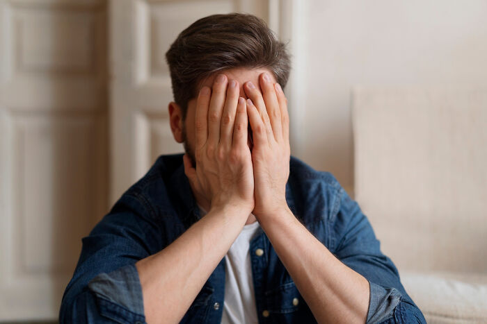 Man covering face with hands, reflecting harsh realities about aging, wearing a denim shirt indoors.