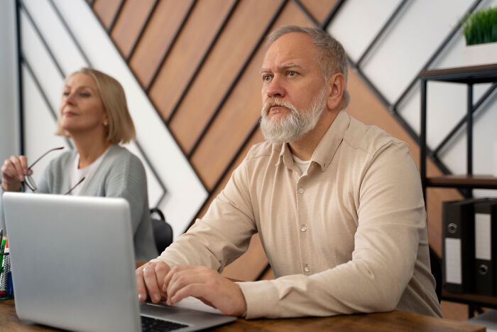 Older man with a beard and woman in a meeting, highlighting aging realities, using a laptop at a wooden desk.